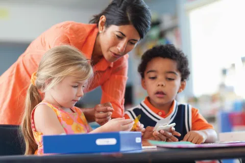 A teacher works with two small children working at their desks.